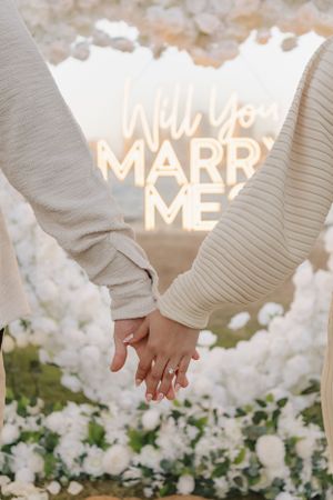 Couple holding hands in front of glowing proposal sign surrounded by white flowers.
