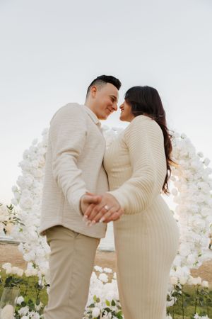 A tender moment between couple under white floral arch during proposal photoshoot.