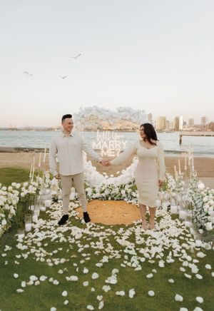 Elegant marriage proposal scene with white floral arch and rose petals scattered on coastal lawn.