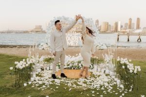 Romantic proposal setup with illuminated arch and white flowers on grass overlooking city skyline at sunset.