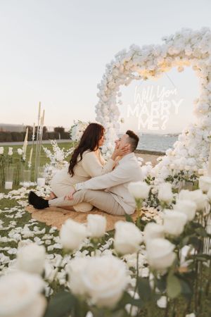 Intimate proposal moment under white floral arch with roses and city view at dusk.