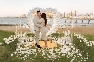 Romantic engagement scene with white floral arch and rose petals against coastal city backdrop.