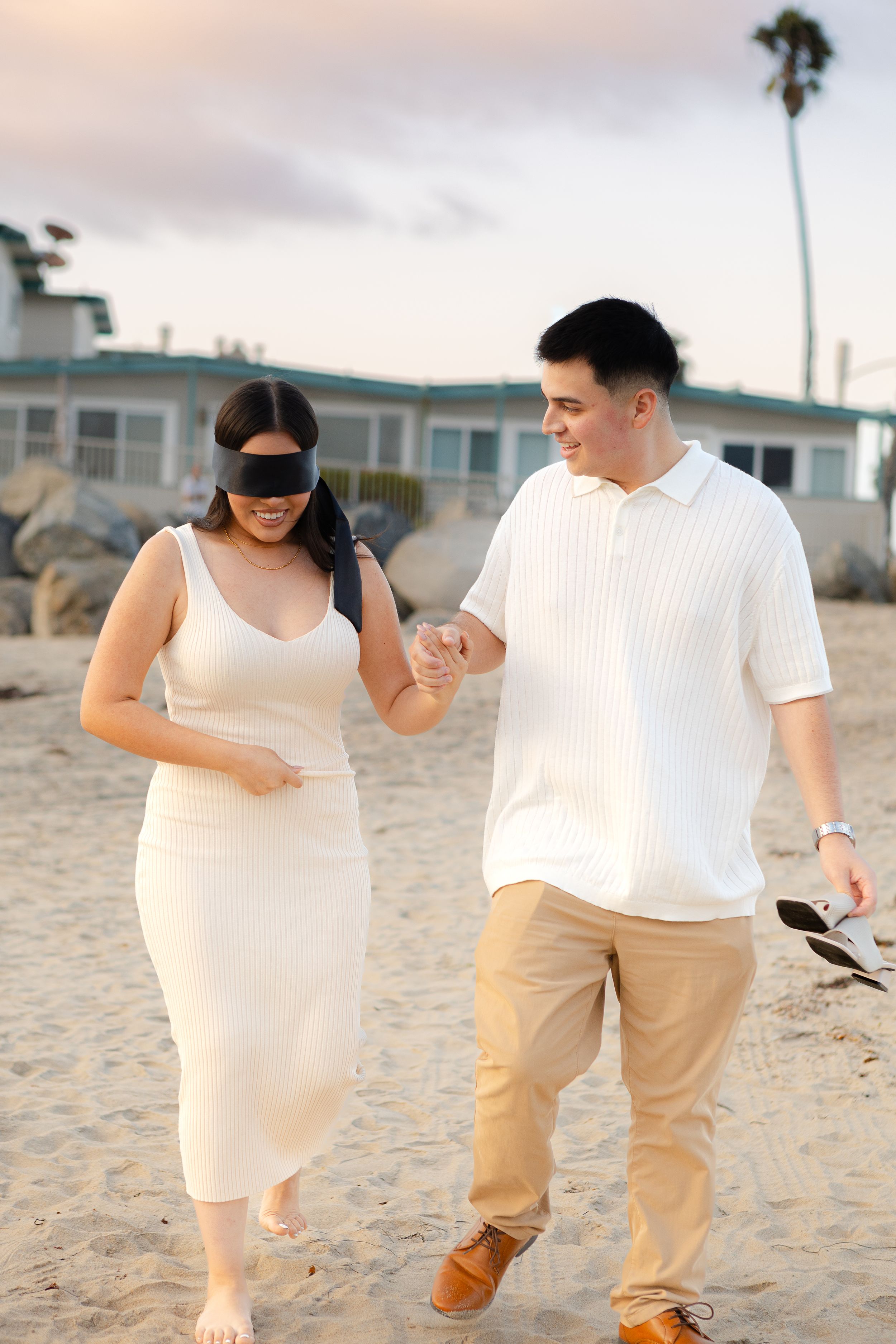 Couple walking together on a sandy beach at sunset near coastal homes and palm trees.