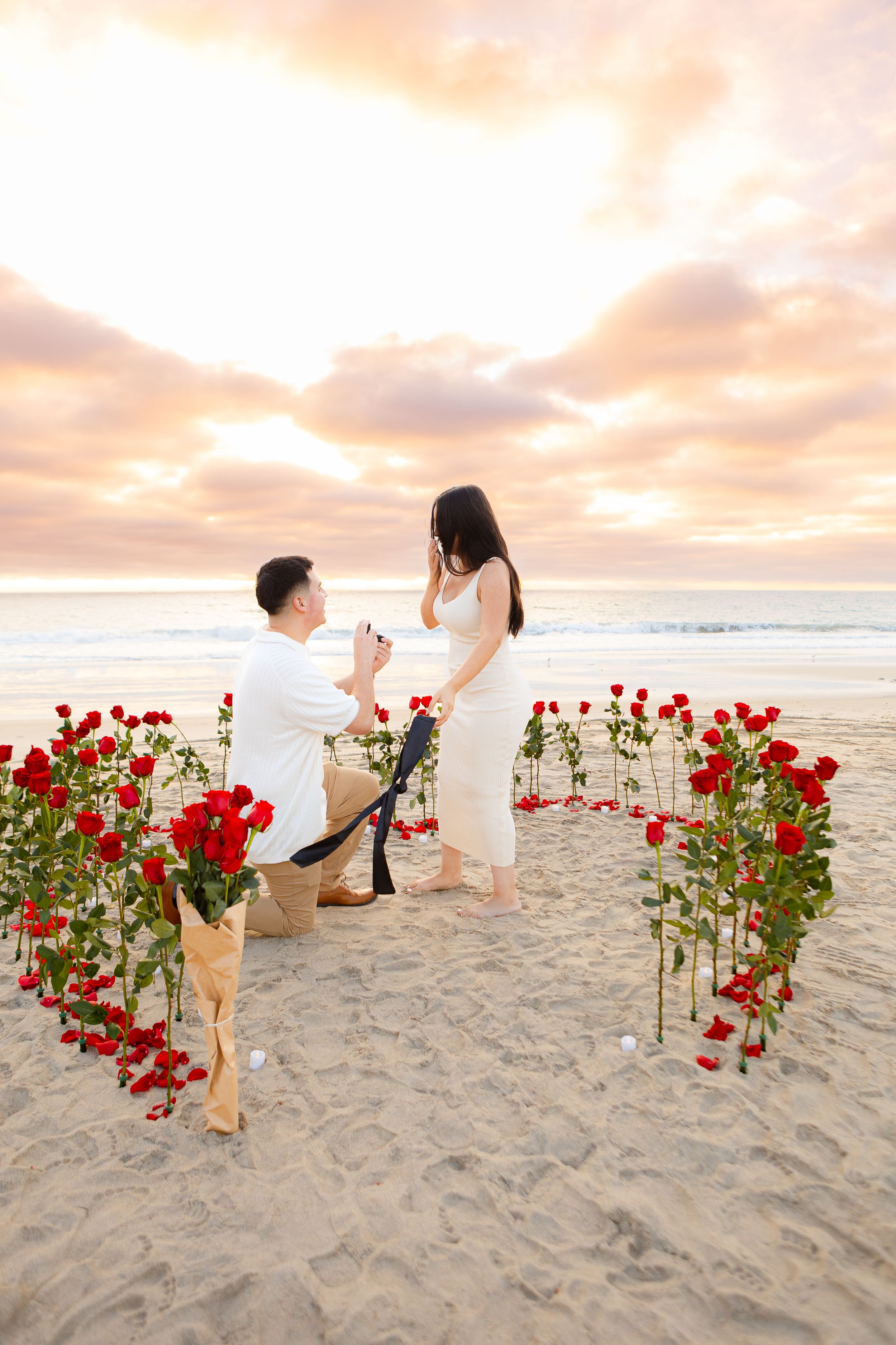 Romantic beach proposal setup with red roses lining a path in the sand during golden hour.