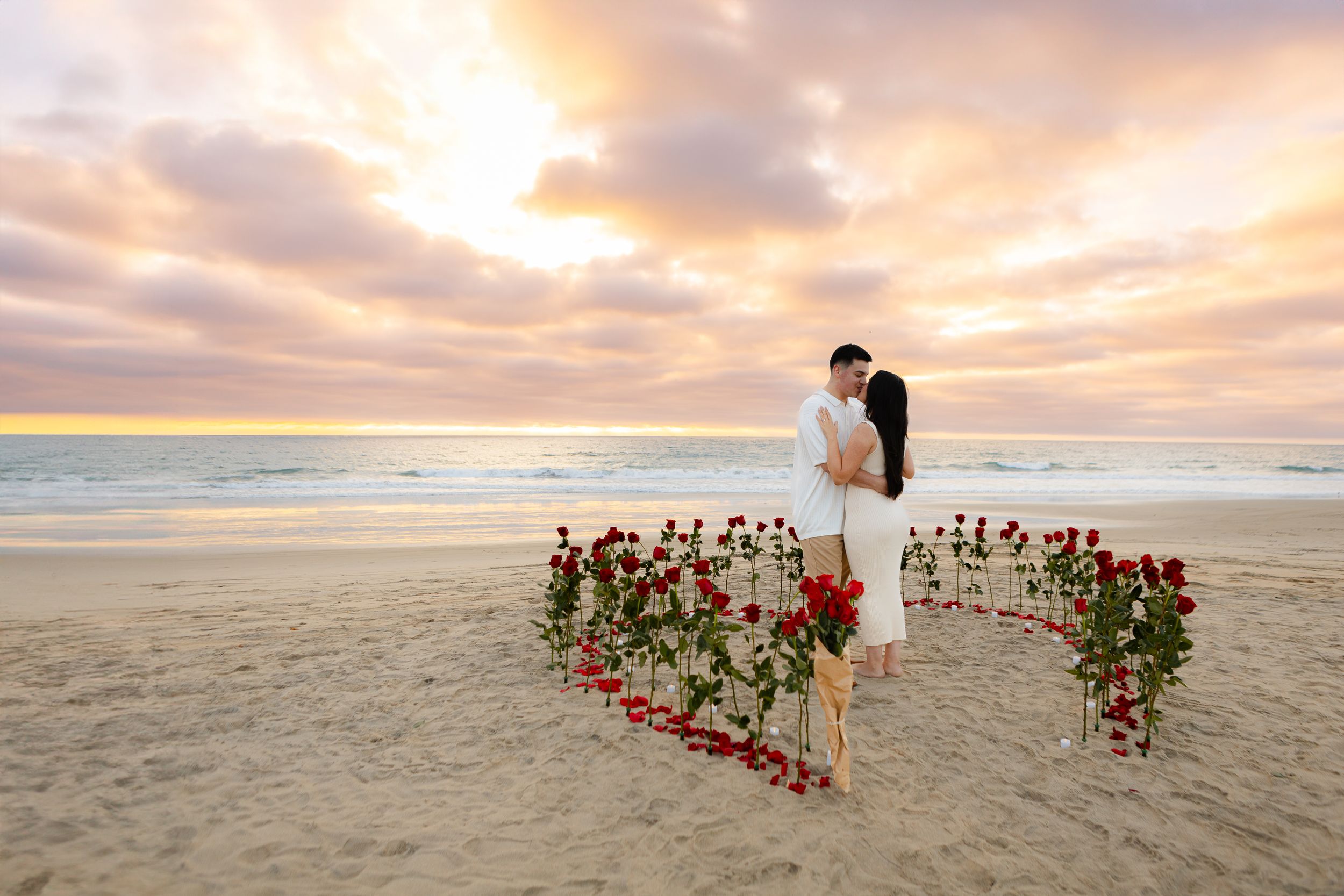 Heart-shaped arrangement of red roses on beach sand with ocean and sunset sky in background.