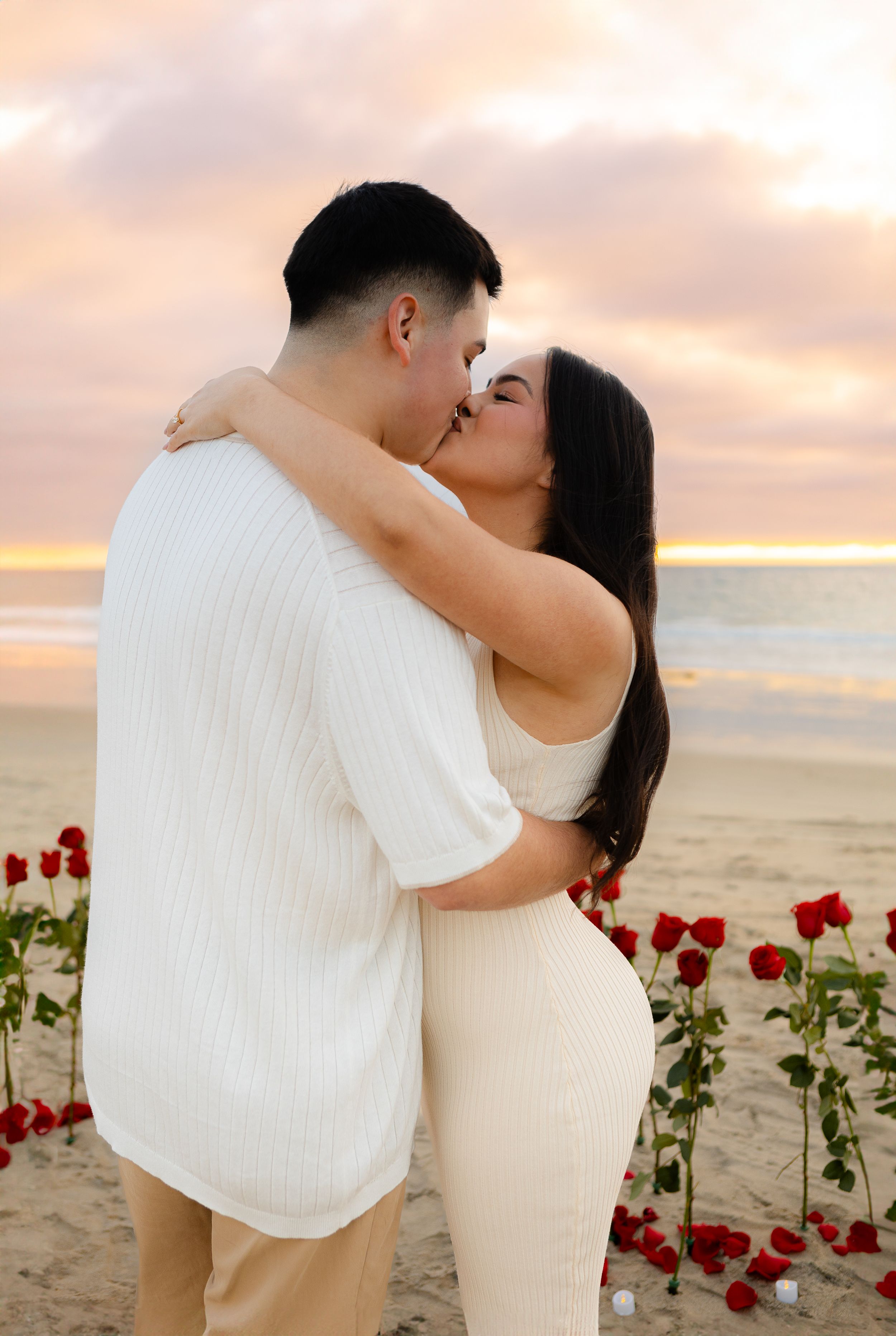 Romantic beach moment with white outfits surrounded by red roses at sunset.