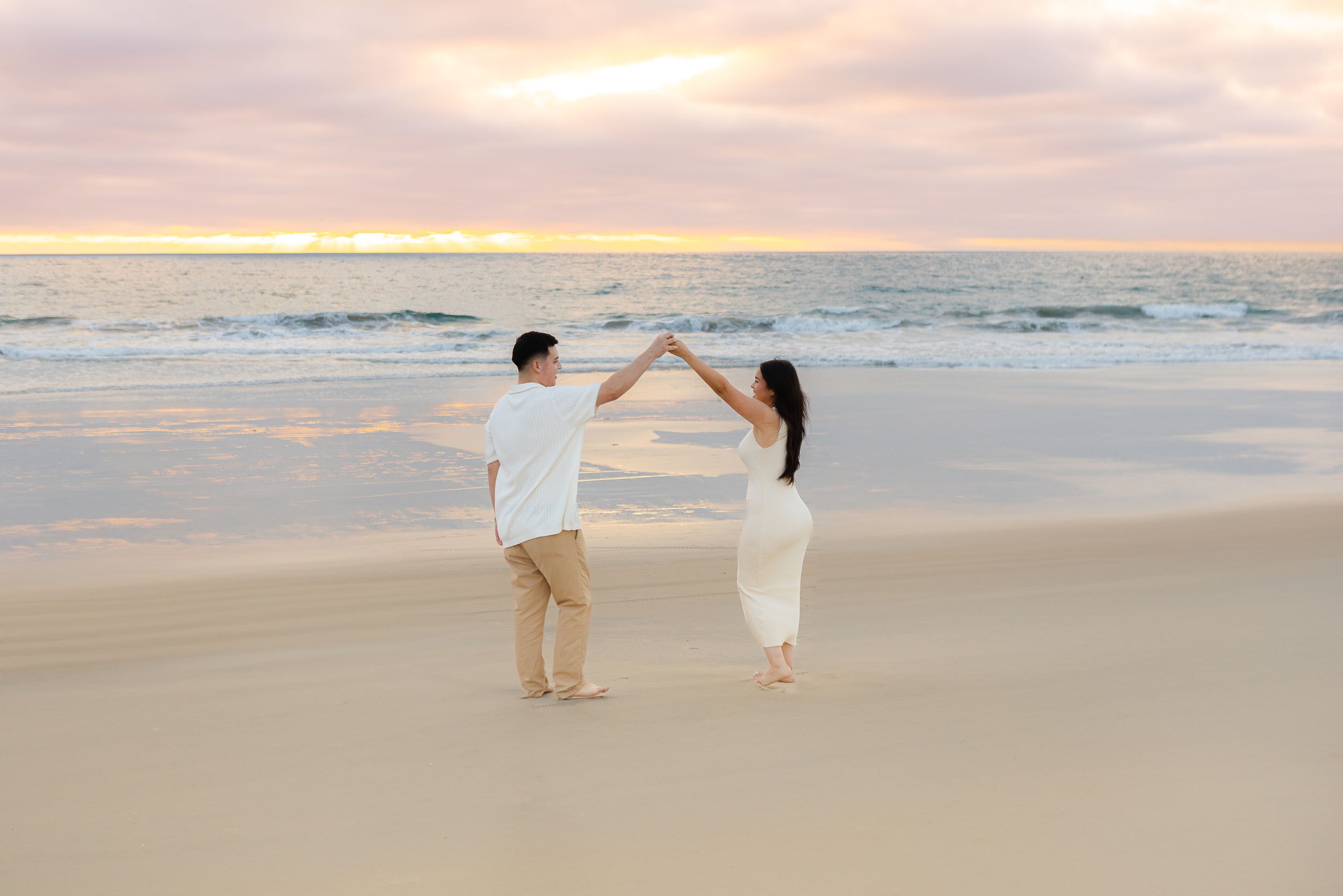Couple in white dancing on beach at sunset with ocean waves in background.
