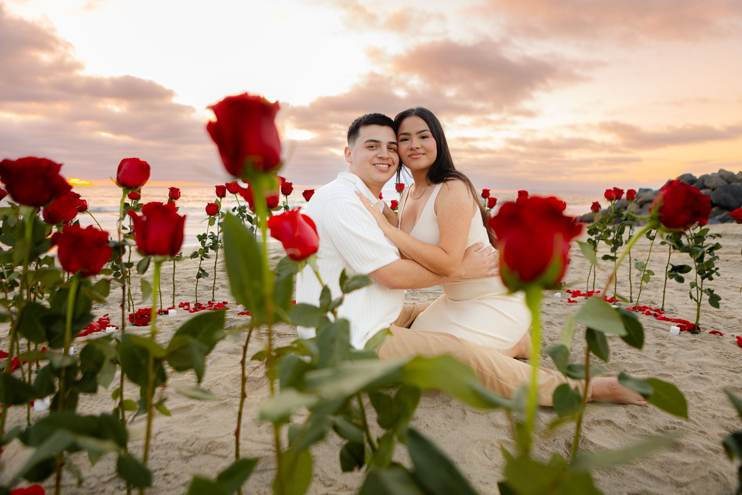 Romantic beach photoshoot with red roses in the foreground during a beautiful sunset.