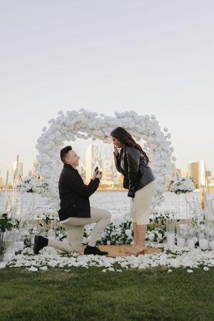 Marriage proposal moment under white floral arch with city skyline in background.
