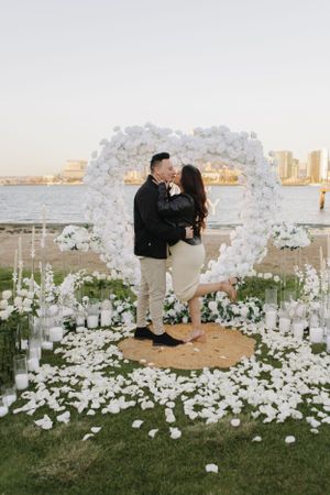 Engagement celebration under white floral arch with rose petals scattered on ground.