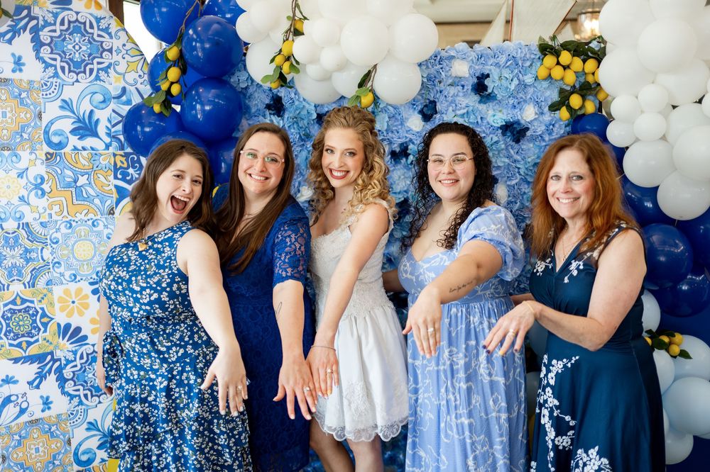A group in coordinated blue and white dresses poses together against a festive balloon arch backdrop.