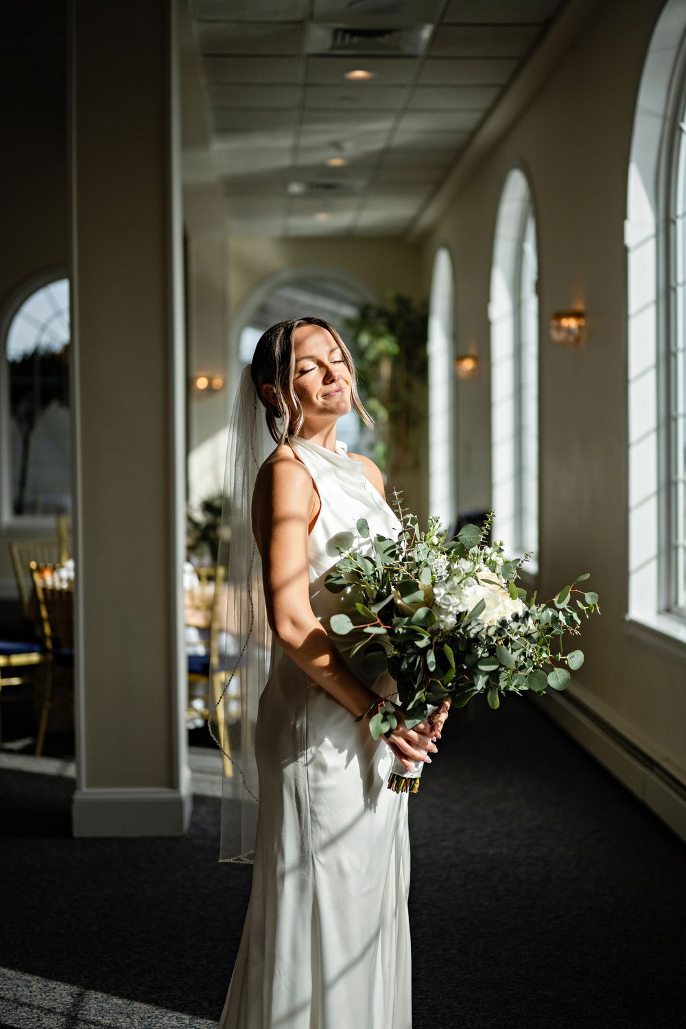 Bride in white gown holding lush greenery bouquet stands in sunlit venue hallway with elegant arched windows.