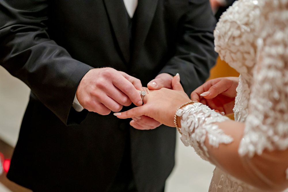 Close-up of hands exchanging wedding rings during an indoor ceremony with black formal attire.