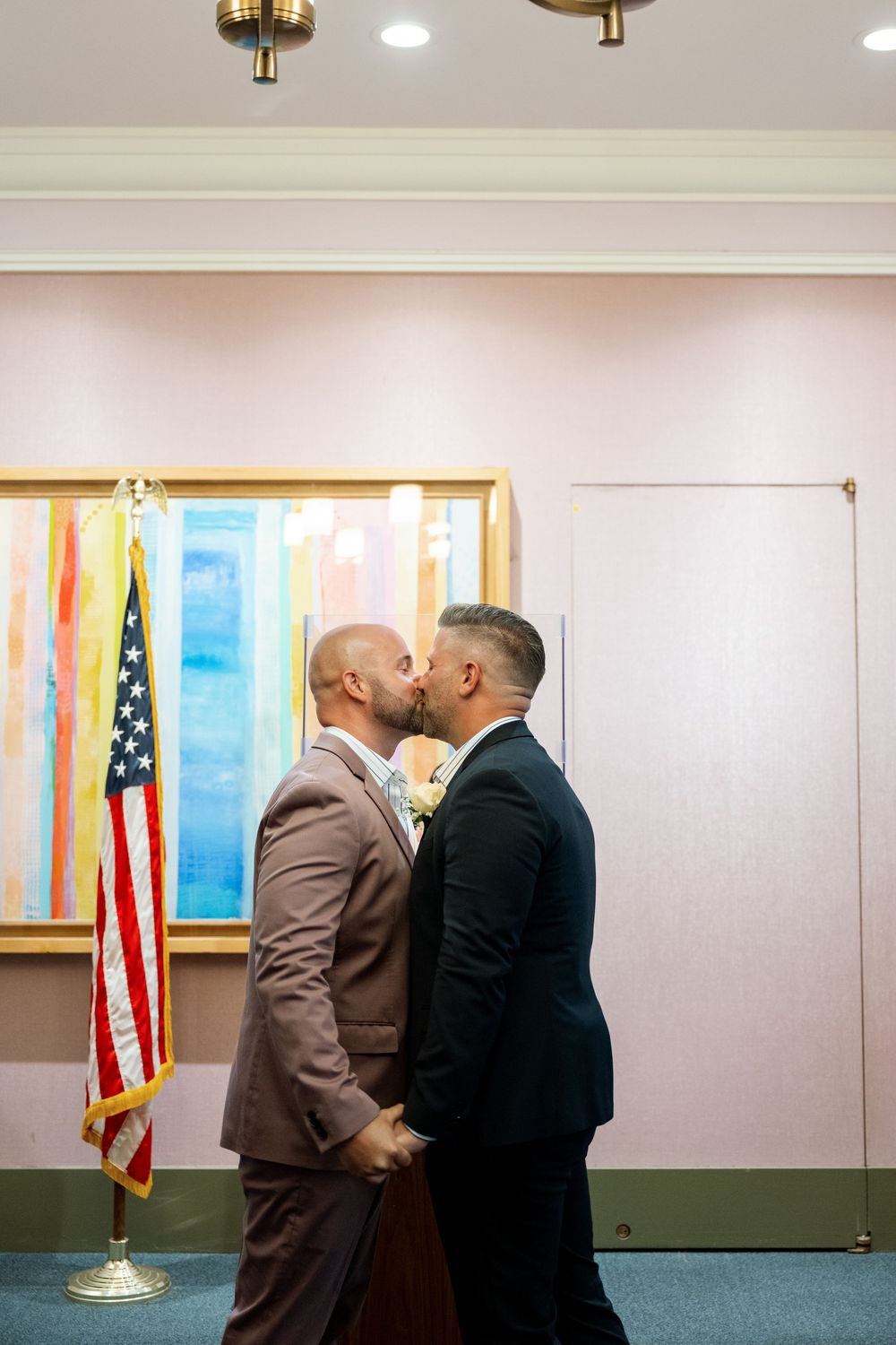 Two people in formal attire stand in an office setting with an American flag and whiteboard visible in the background.