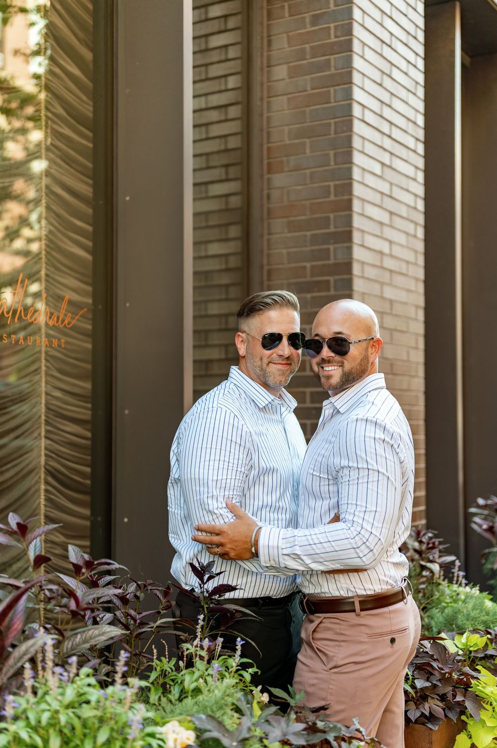 Two people in matching striped shirts and khaki pants embrace and laugh together in front of a brick building.