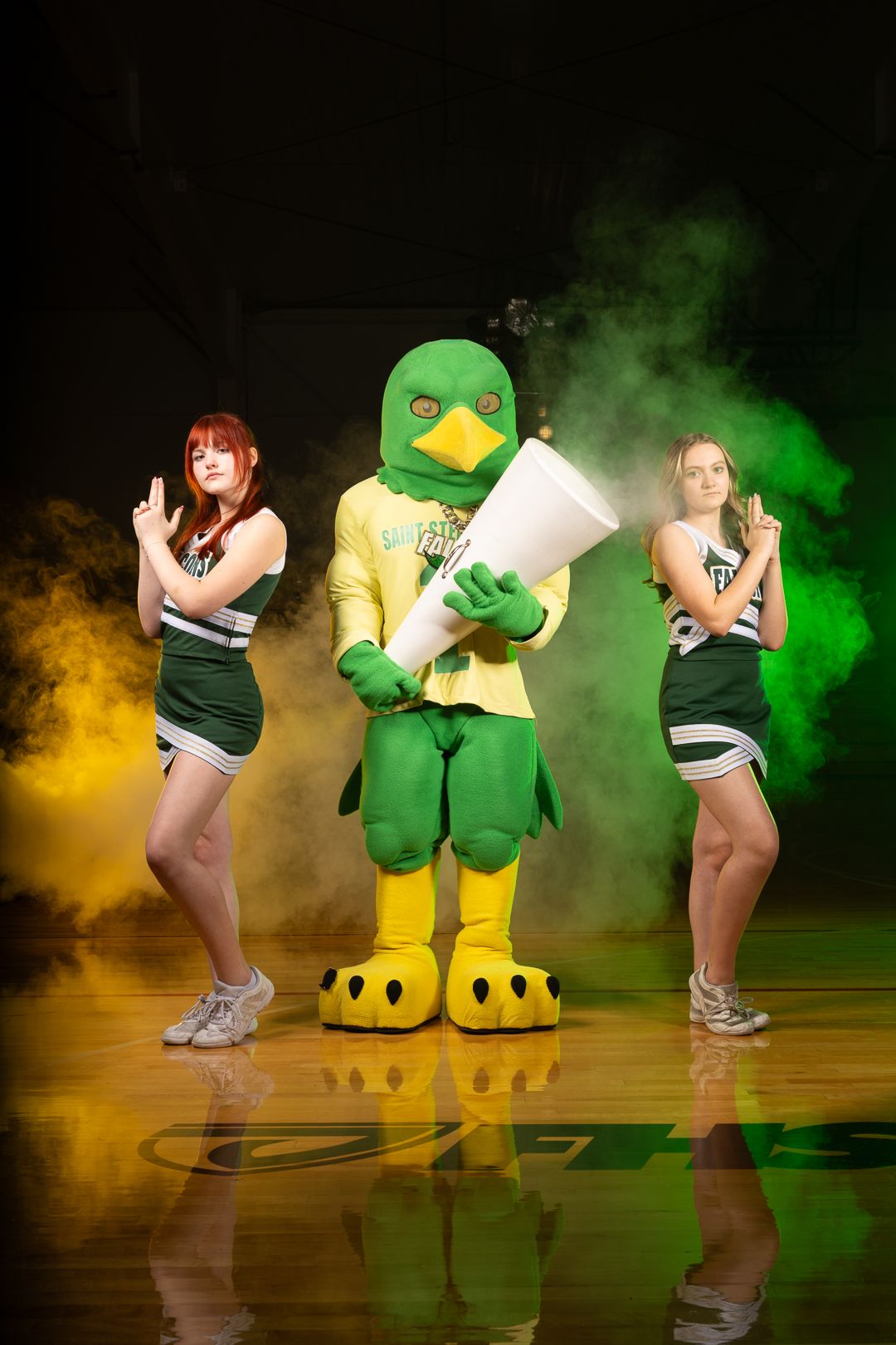 School mascot in green duck costume poses with cheerleaders amid colored smoke on basketball court.