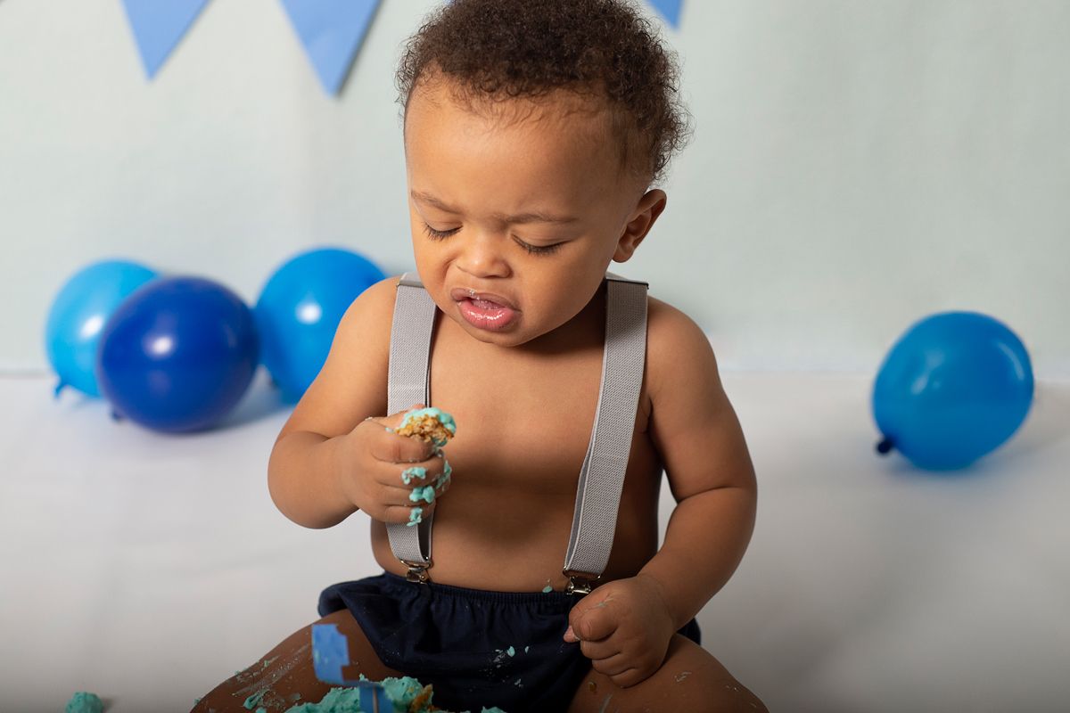 toddler boy in suspenders looks a his hands with the icing from his cake during his first birthday cake smash session, with blue balloons