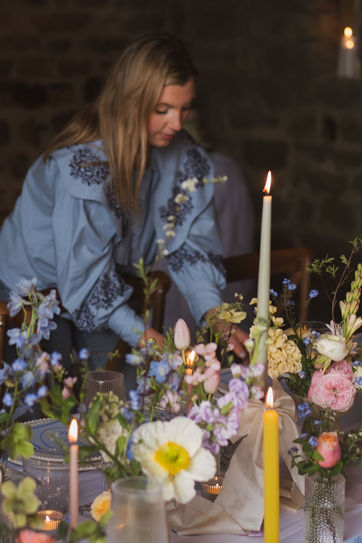 A romantic table setting with pink and yellow flowers, lit candles and someone in a blue embroidered dress arranging the decor.