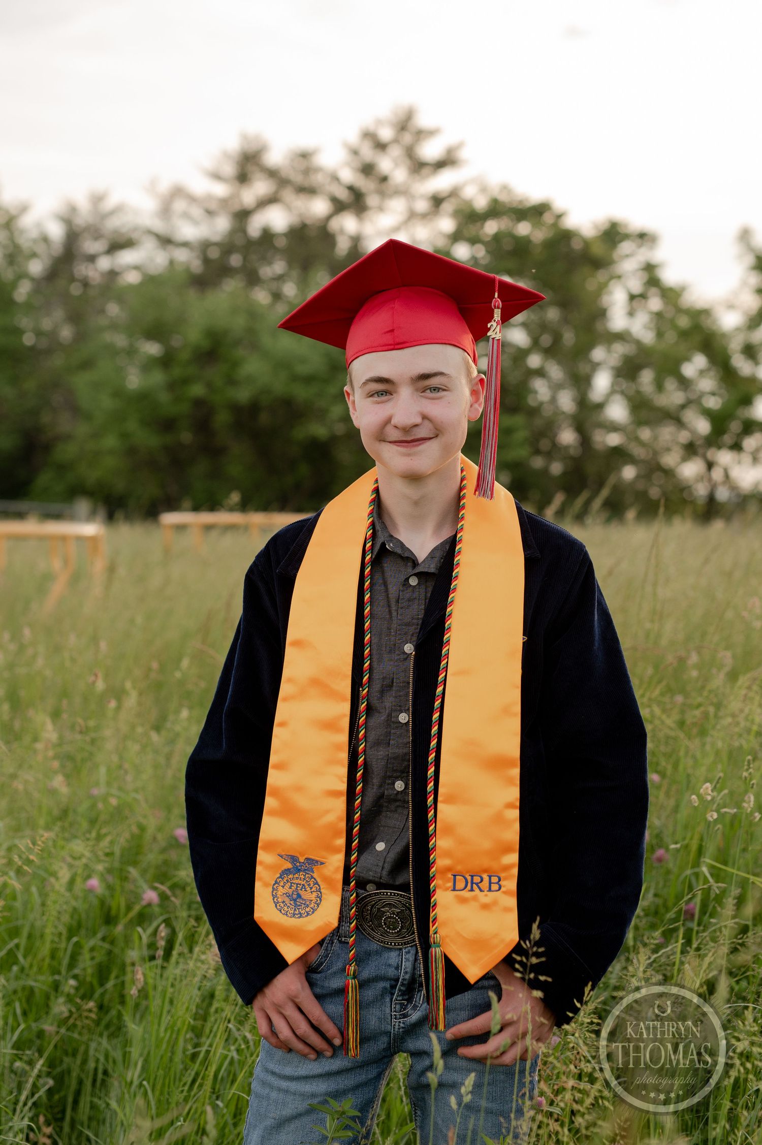 Graduate in red cap and gold stole stands proudly in FFA jacket amidst tall grass field.