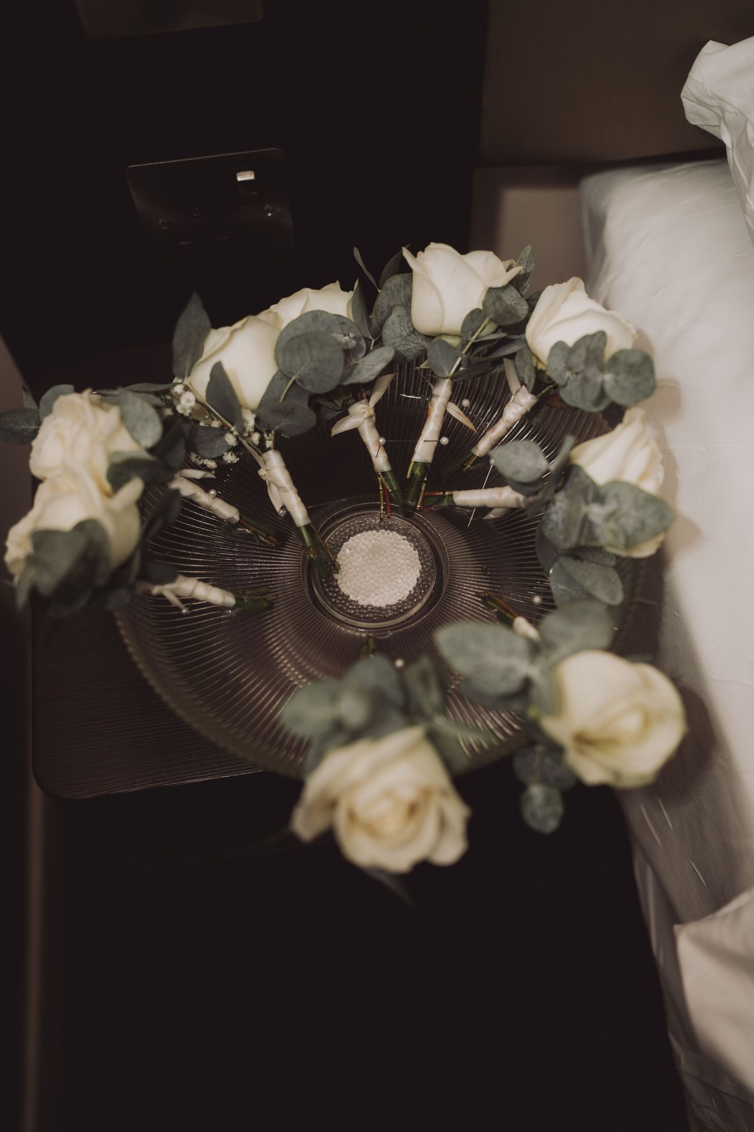 White roses and eucalyptus arranged in a circular pattern on a dark surface creating a wedding bouquet display.