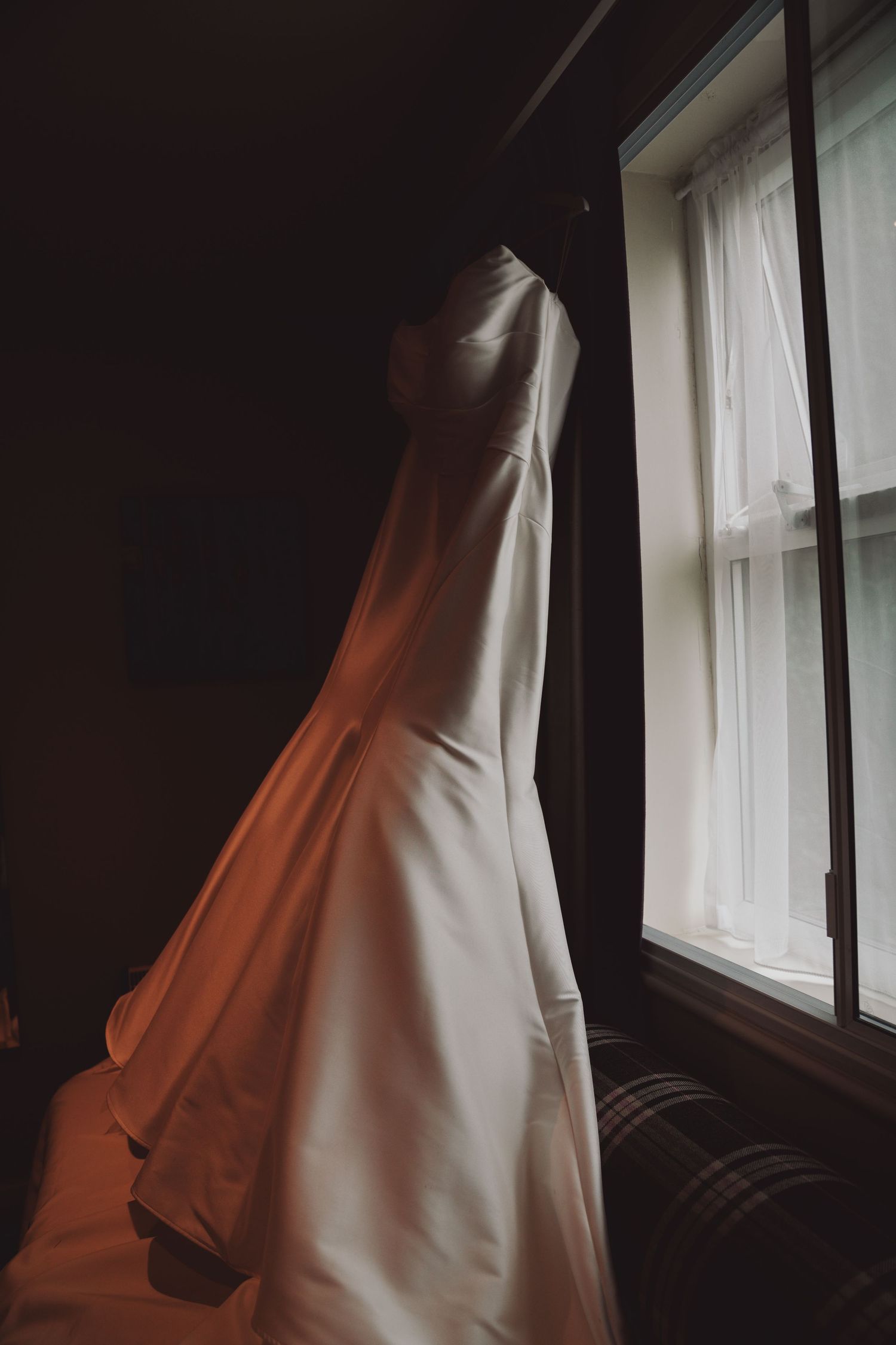 An elegant white wedding dress hangs by a window in dramatic lighting creating shadows and silhouettes.