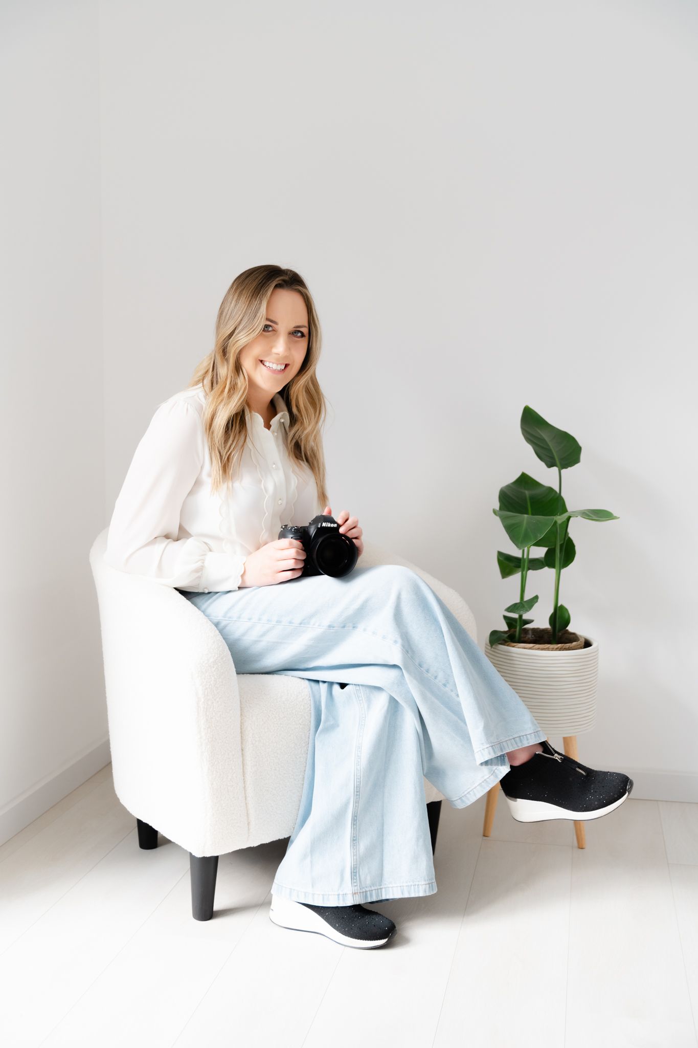 A person in a white sweater and light blue jeans sits in a cream-colored chair next to a potted plant in a minimal setting.
