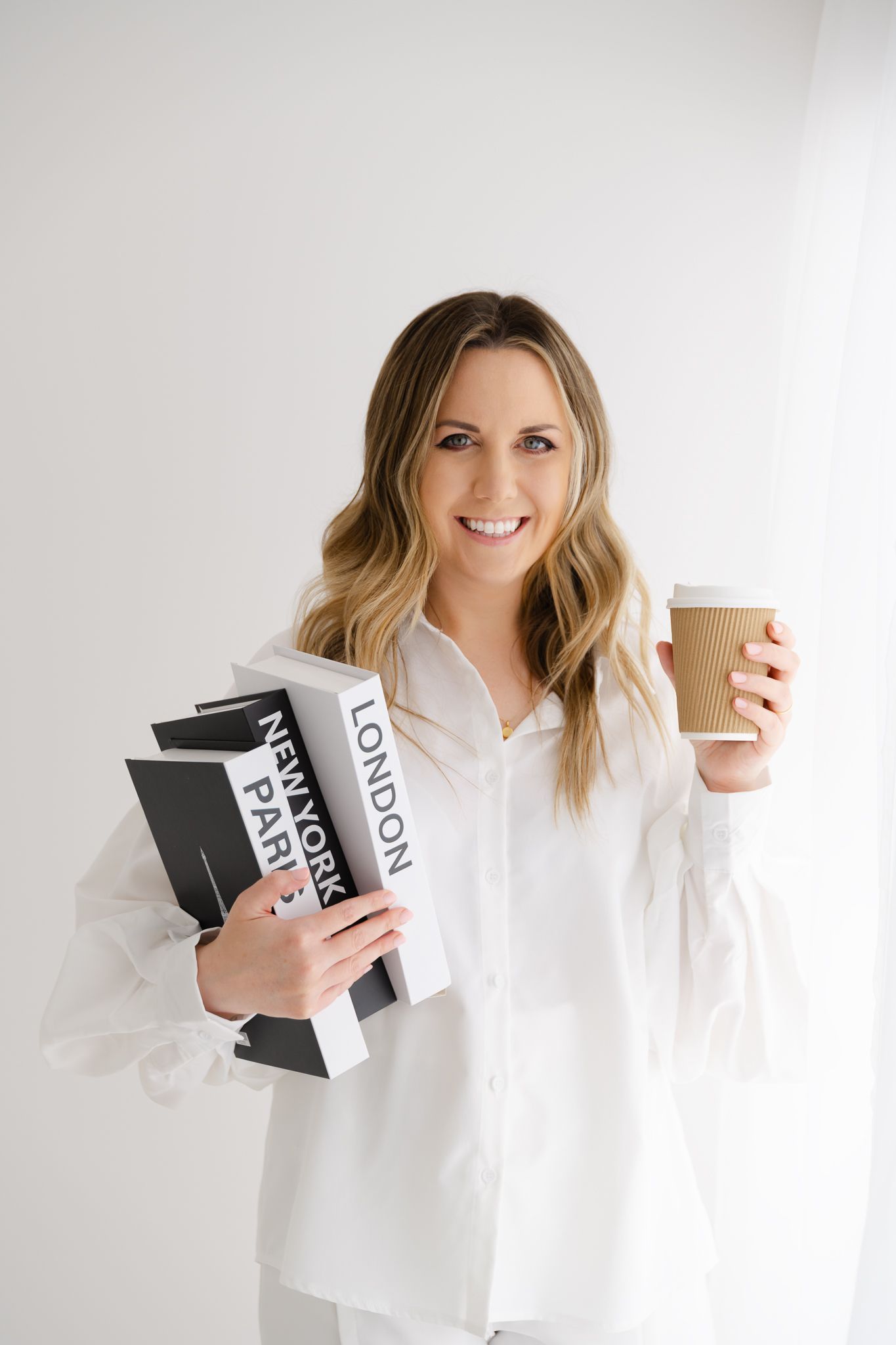 Professional in white outfit holds business folders and coffee cup against white wall.