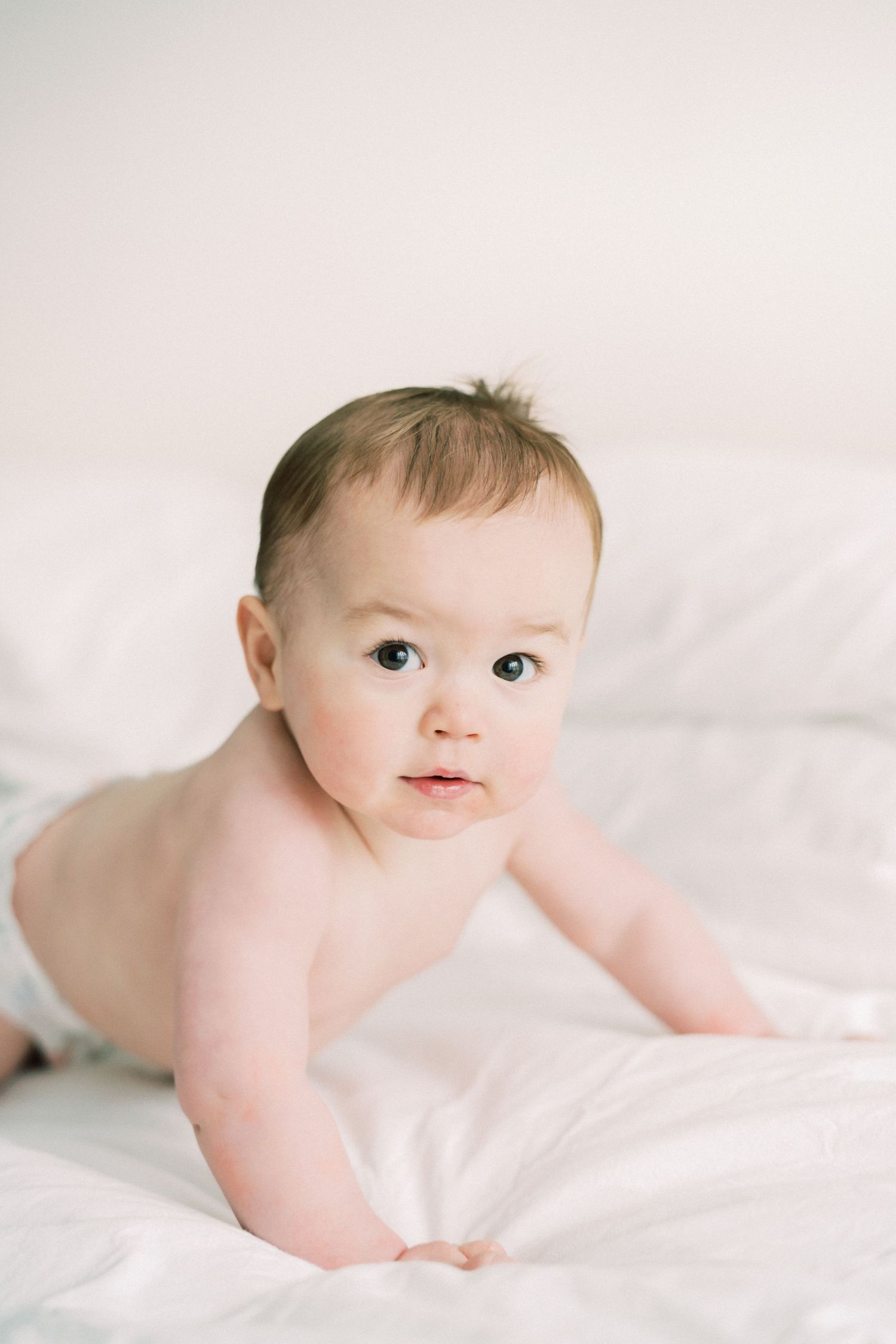 A baby wearing a white lace dress and bow headband crawls on a white backdrop with roses in the background.