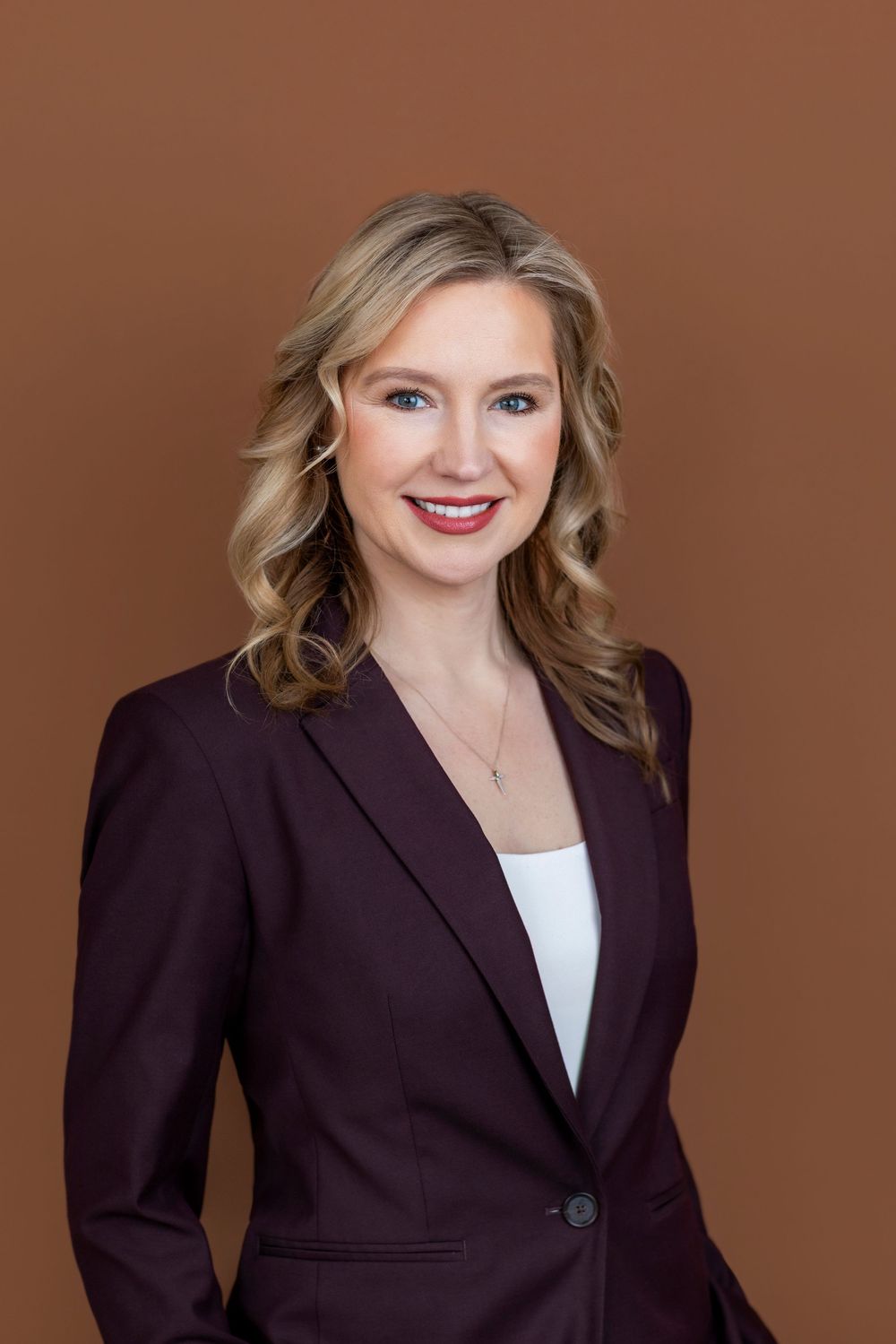 Professional woman in dark blazer smiling confidently against brown background, ideal corporate headshot.
