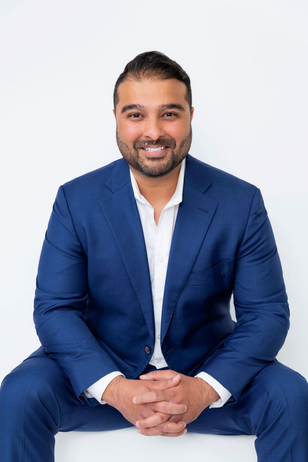 Professional wearing a navy blue suit and white shirt sits casually in various poses against a light background.