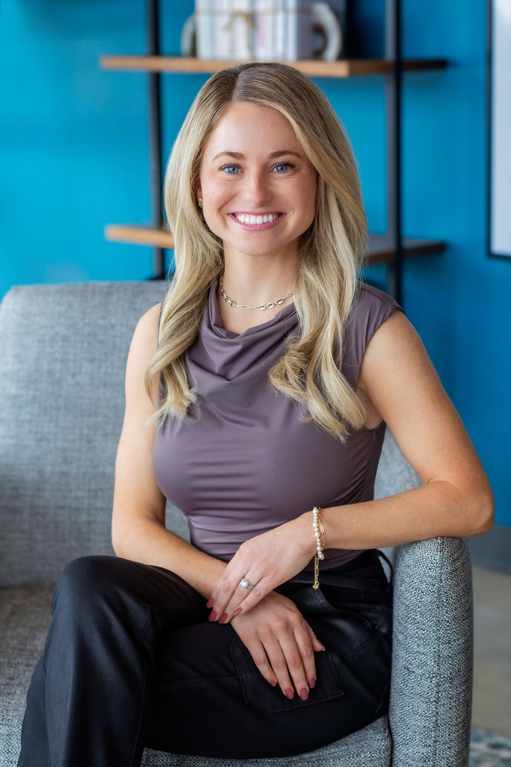 Professional headshot of someone in a grey sleeveless top and black pants sitting on a grey chair against a bright blue wall.