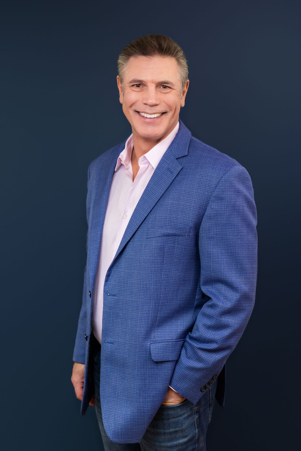 Professional headshot of a person in a blue blazer and pink shirt smiling against a dark background.