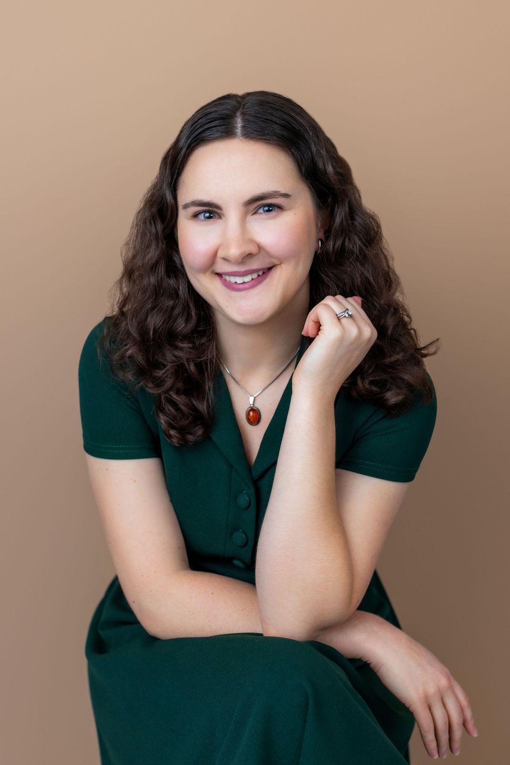 Portrait of someone in an emerald green dress with curly dark hair and a pendant necklace against a beige background.