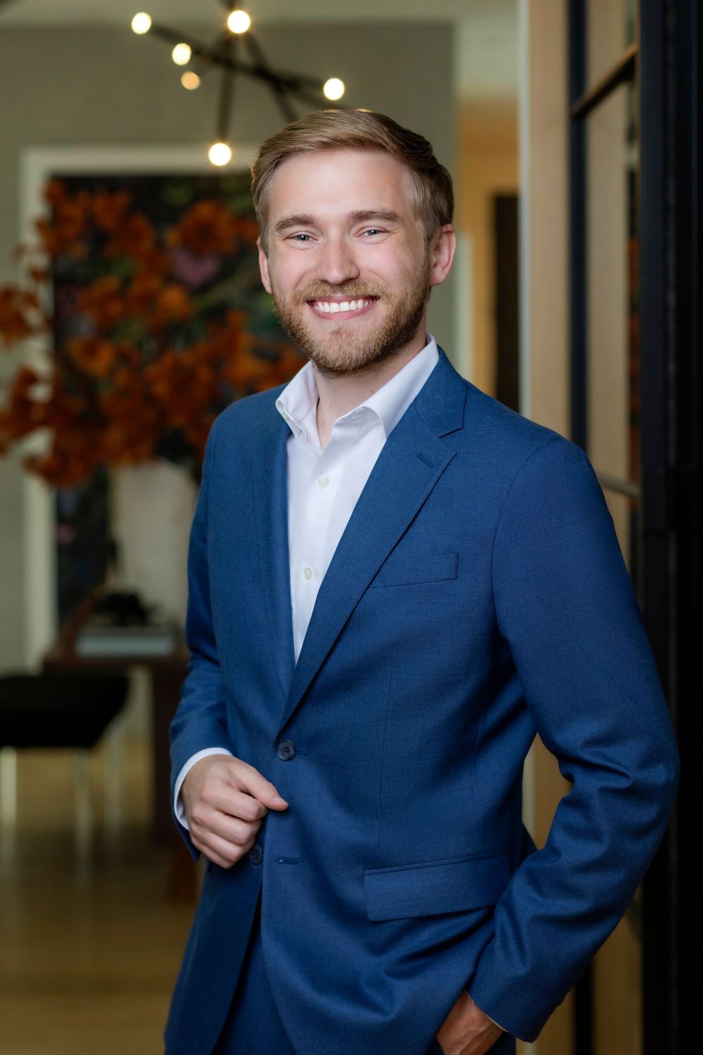 Professional portrait of someone in a navy blue suit and white dress shirt standing in an office with bokeh lighting.