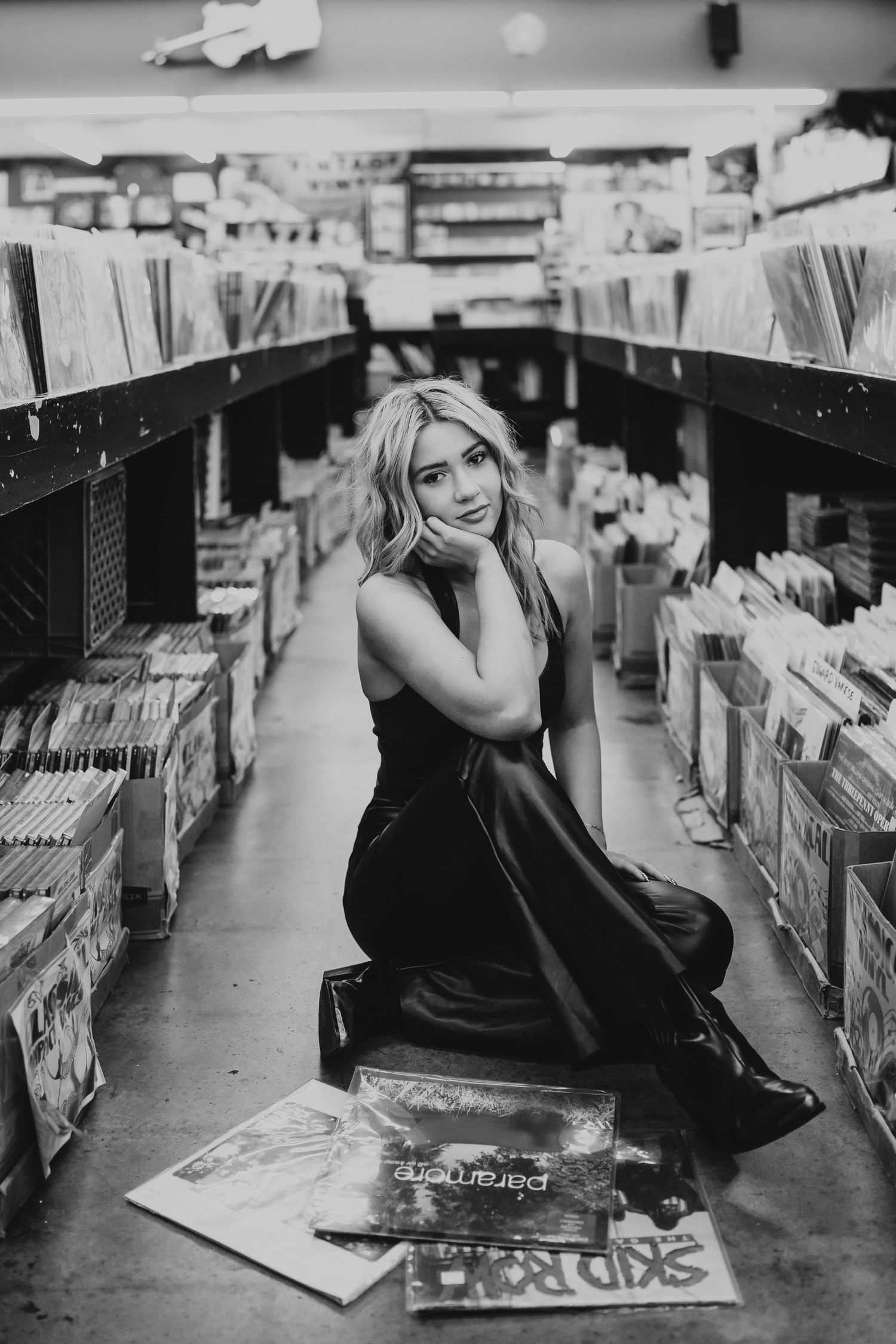 A woman sitting on the floor of a record store, surrounded by vinyl records, looking through a collection with a smile.