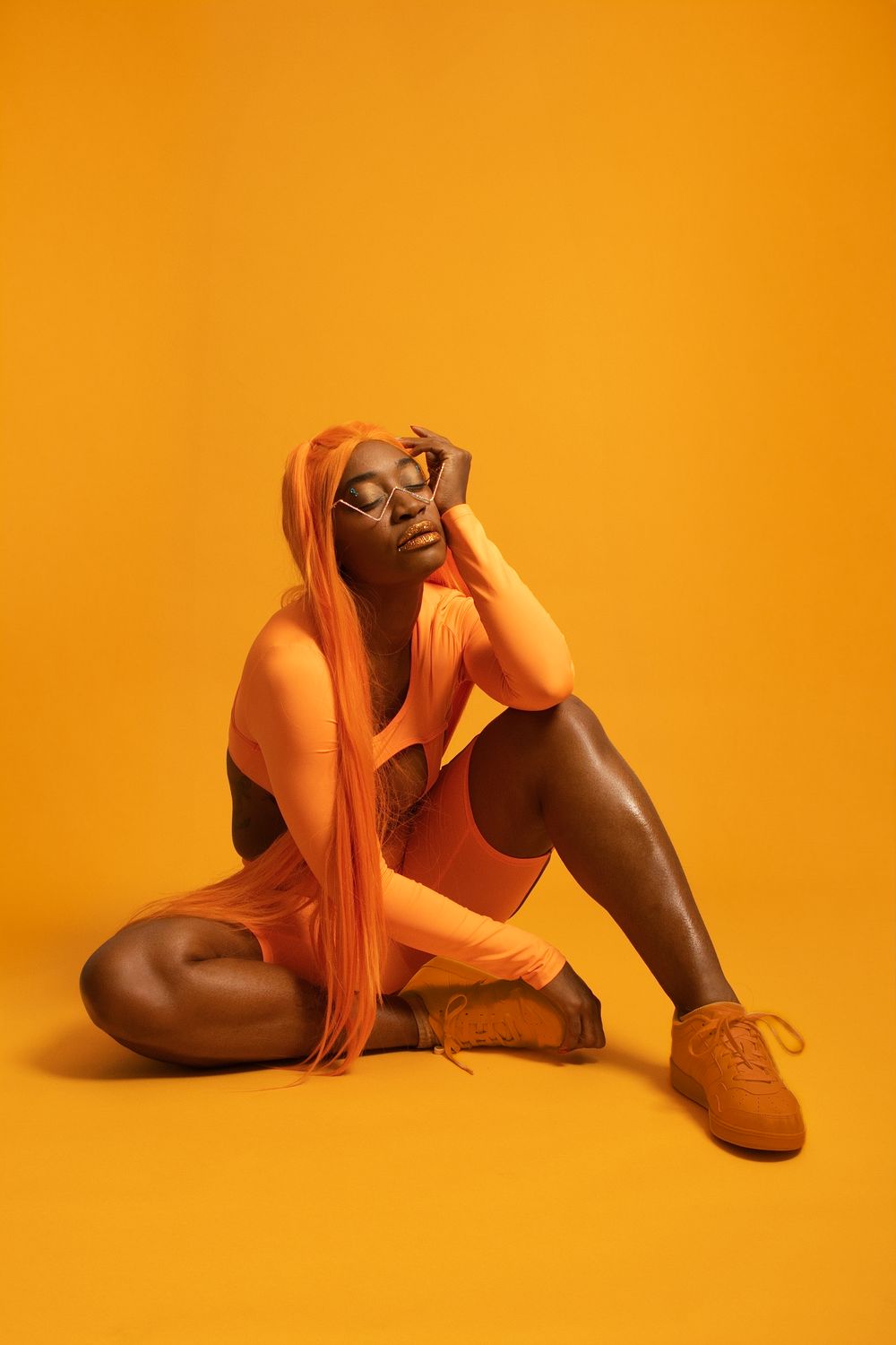 Black woman in all-orange outfit with long orange hair sitting on yellow background in a fashion studio shoot.