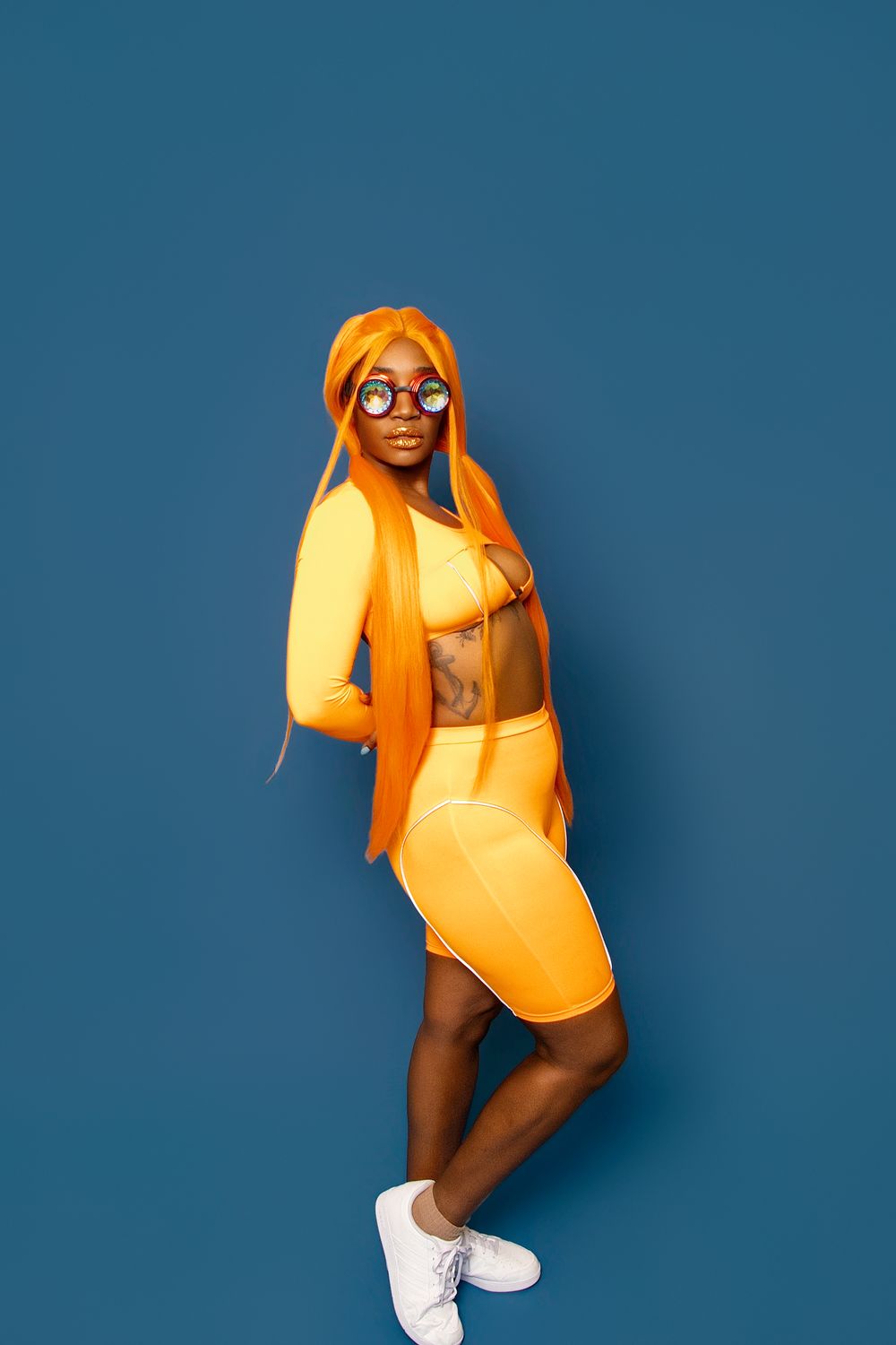 Black woman in vibrant orange outfit and wig with round sunglasses posing against blue background.