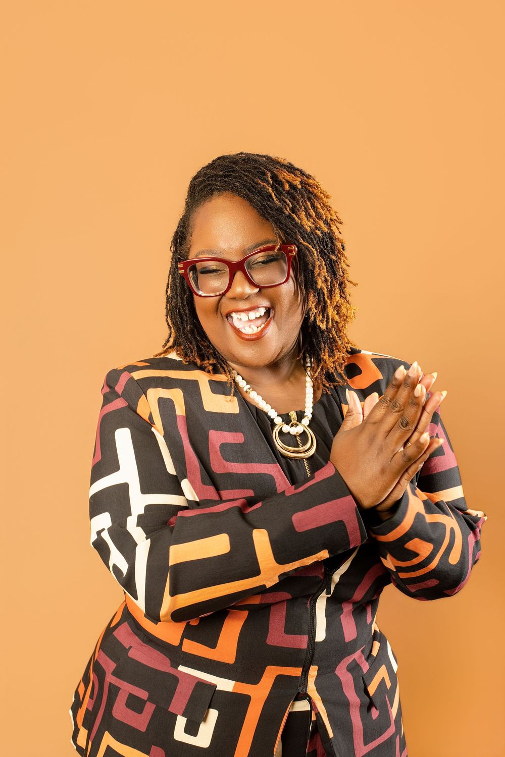 Smiling woman in colorful geometric print blazer, red glasses, pearl necklace, clapping hands against orange background.