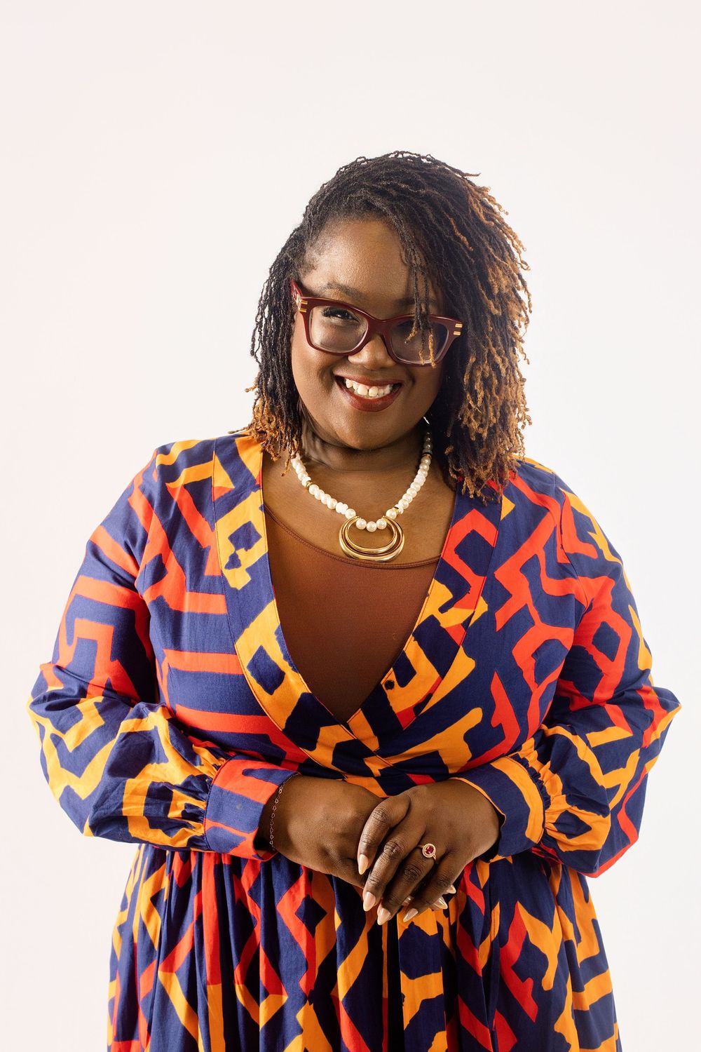 Smiling woman in colorful geometric print dress wearing glasses, pearl necklace, and gold pendant against white background.