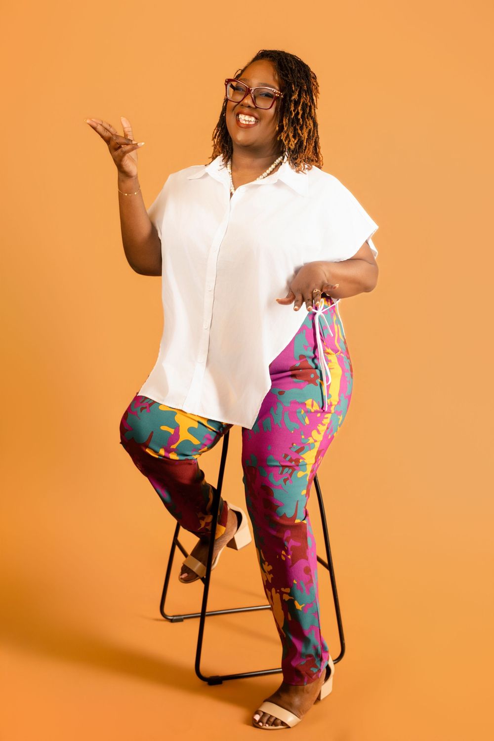 Woman in white blouse and colorful floral pants sitting on stool, smiling, wearing glasses, orange background.