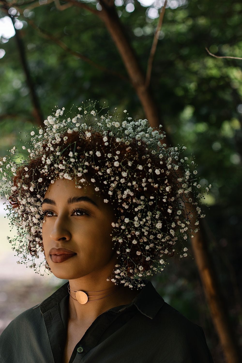 Woman with curly hair covered in delicate baby's breath flowers stands beneath trees, wearing a dark collared shirt and a gold choker.