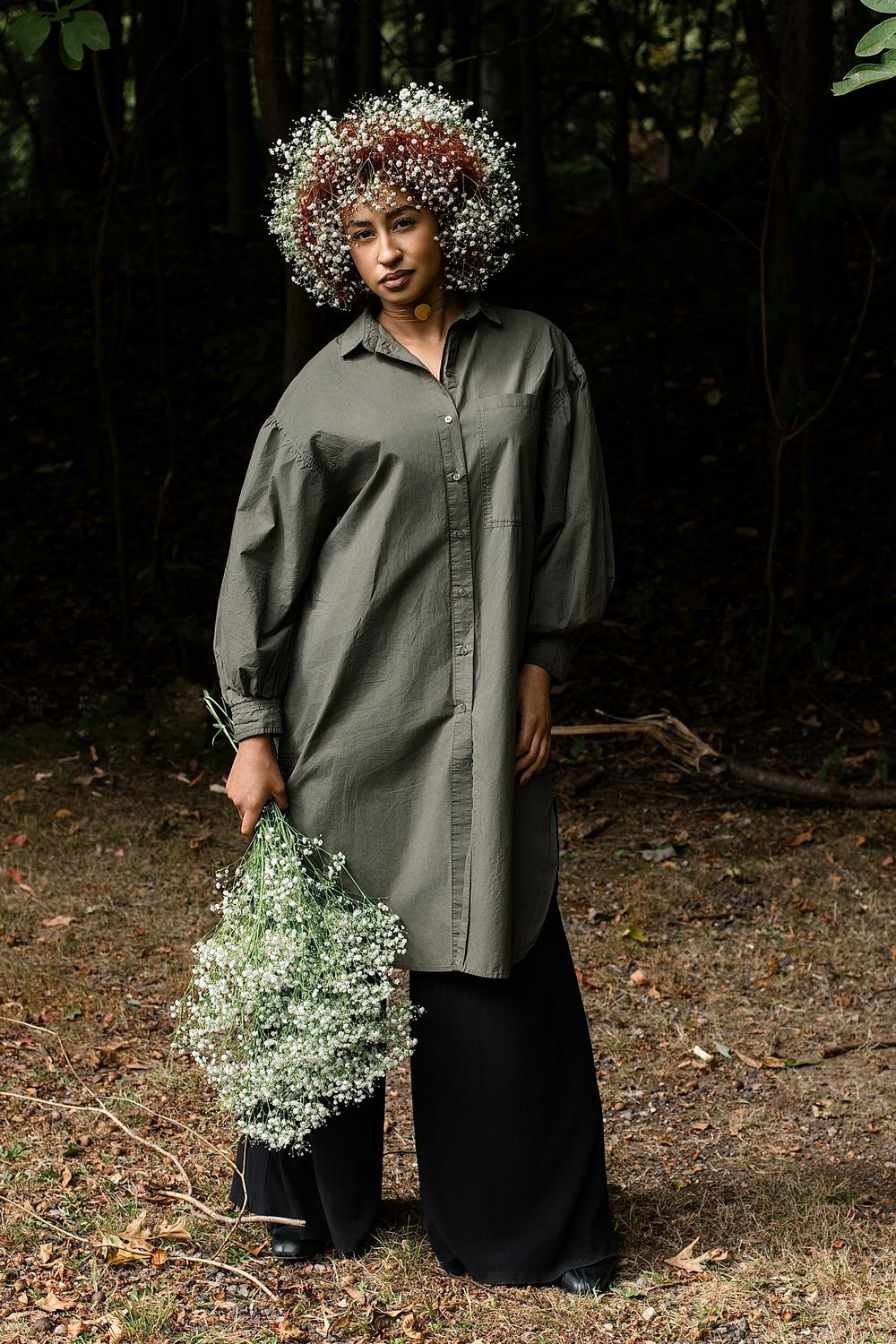Woman wearing a baby's breath flower crown, an olive green oversized shirt dress and black pants, holding a bouquet in a dark forest.