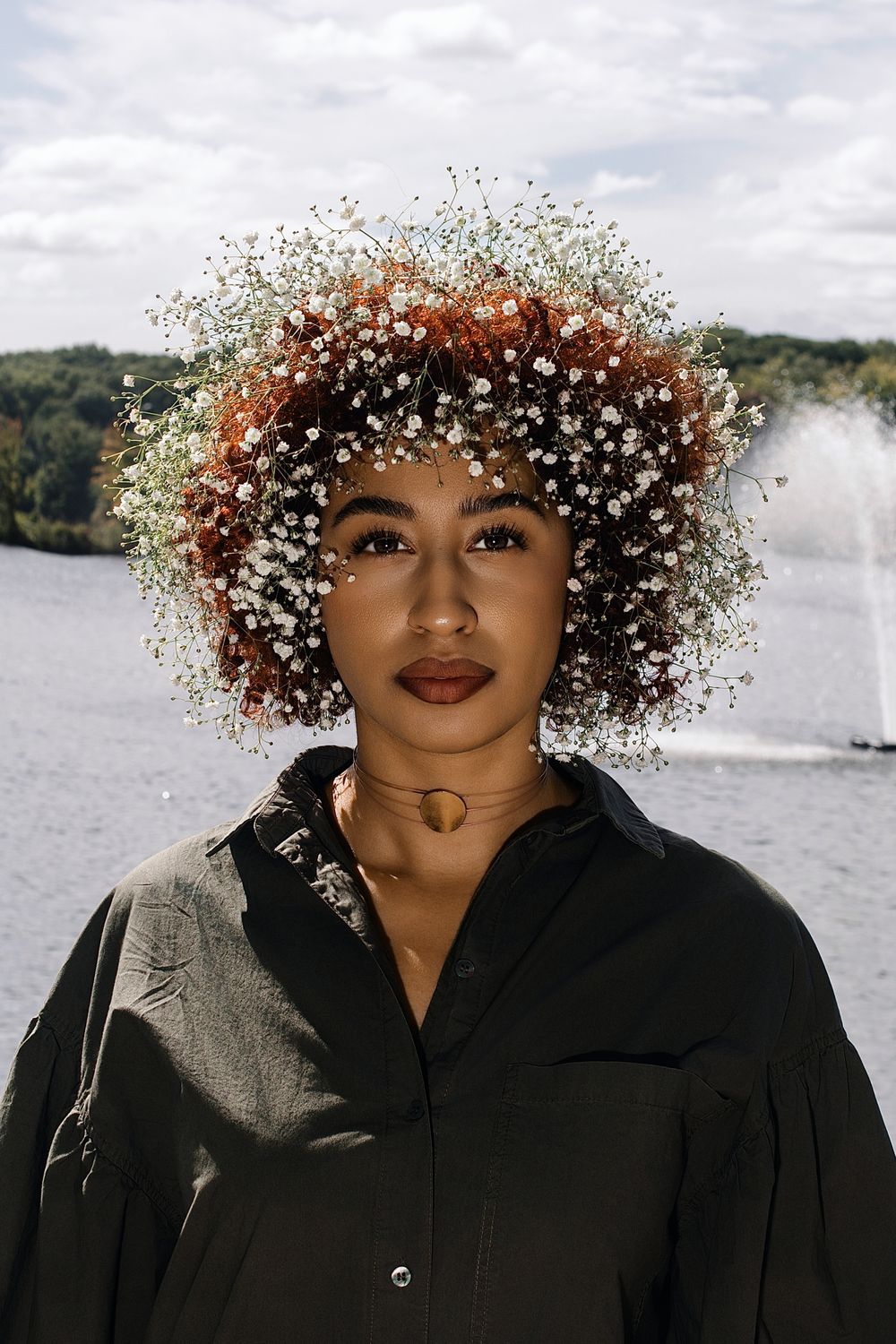 Woman with curly auburn hair covered in baby's breath flowers wears a black button-up shirt and gold necklace beside a lake with a fountain.