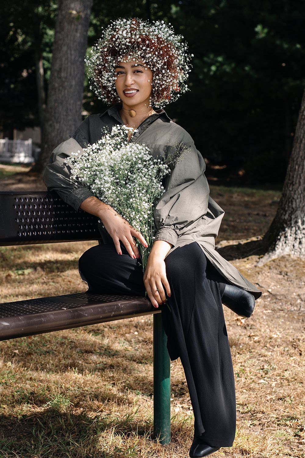 Woman with a baby's breath flower crown smiles while sitting on a park bench holding a bouquet, wearing an olive jacket and black pants.