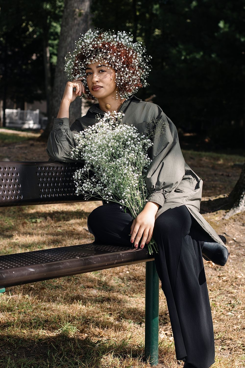 Woman wearing a baby's breath flower crown sits on a park bench holding a bouquet of white gypsophila flowers in dappled sunlight.