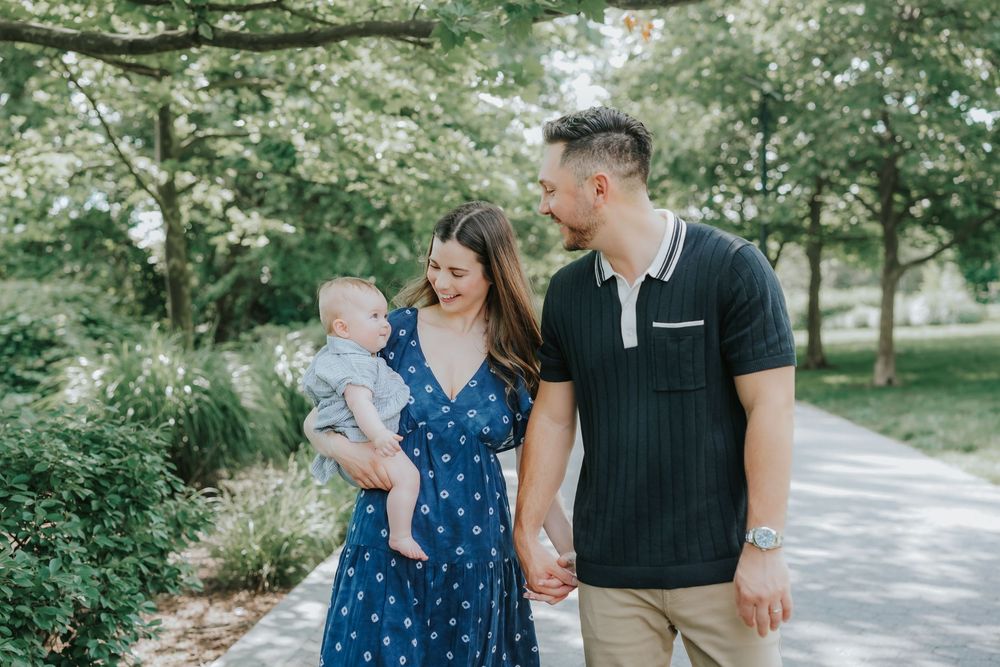 A happy young family with a baby strolls along a park path surrounded by lush green trees on a sunny day.