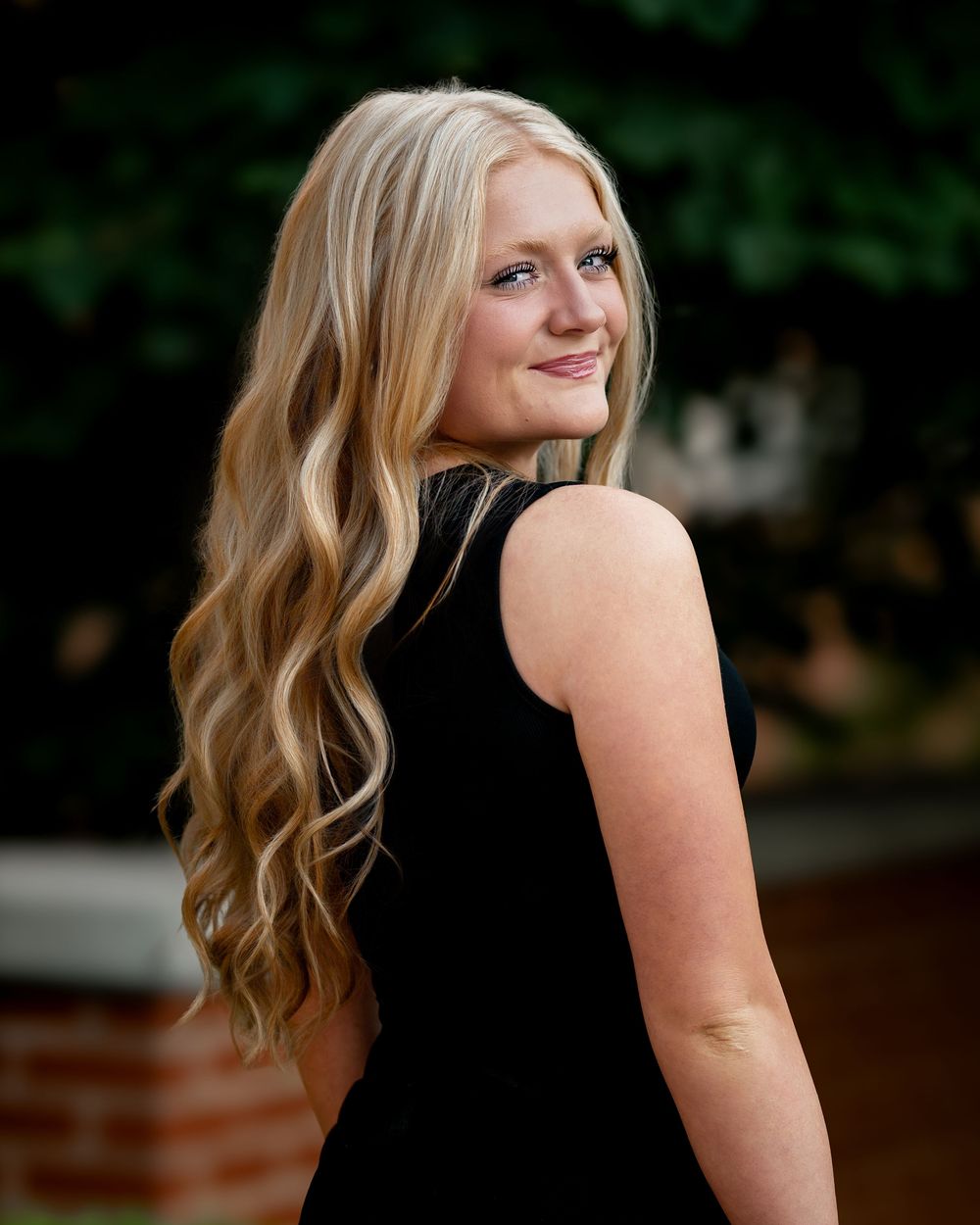 Young blonde woman in black dress smiling over shoulder outdoors, long wavy hair, natural bokeh background. Jen Welker, Kansas City Senior P
