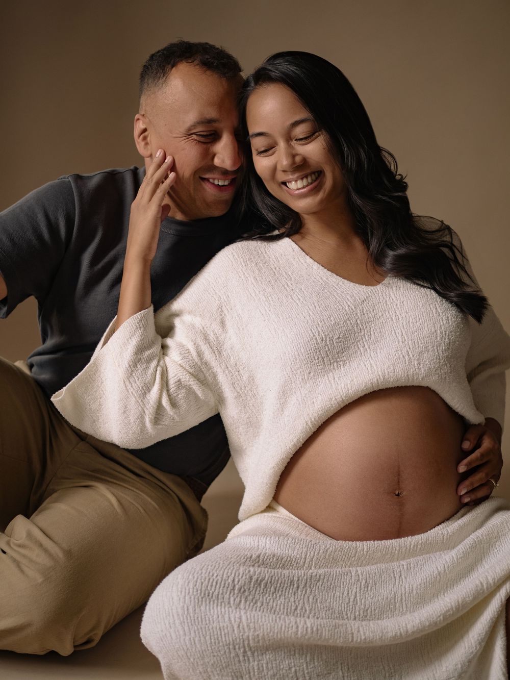 Intimate maternity photo series showing couple in warm embrace against neutral backdrop wearing white sweater.