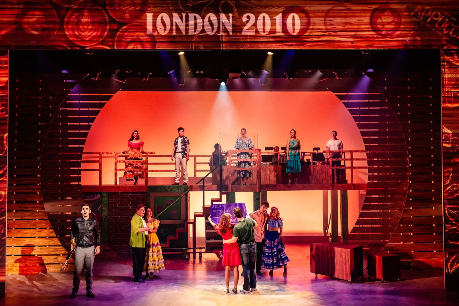 Theater stage with red lighting displays London 2010 text above performers on a multi-level set.