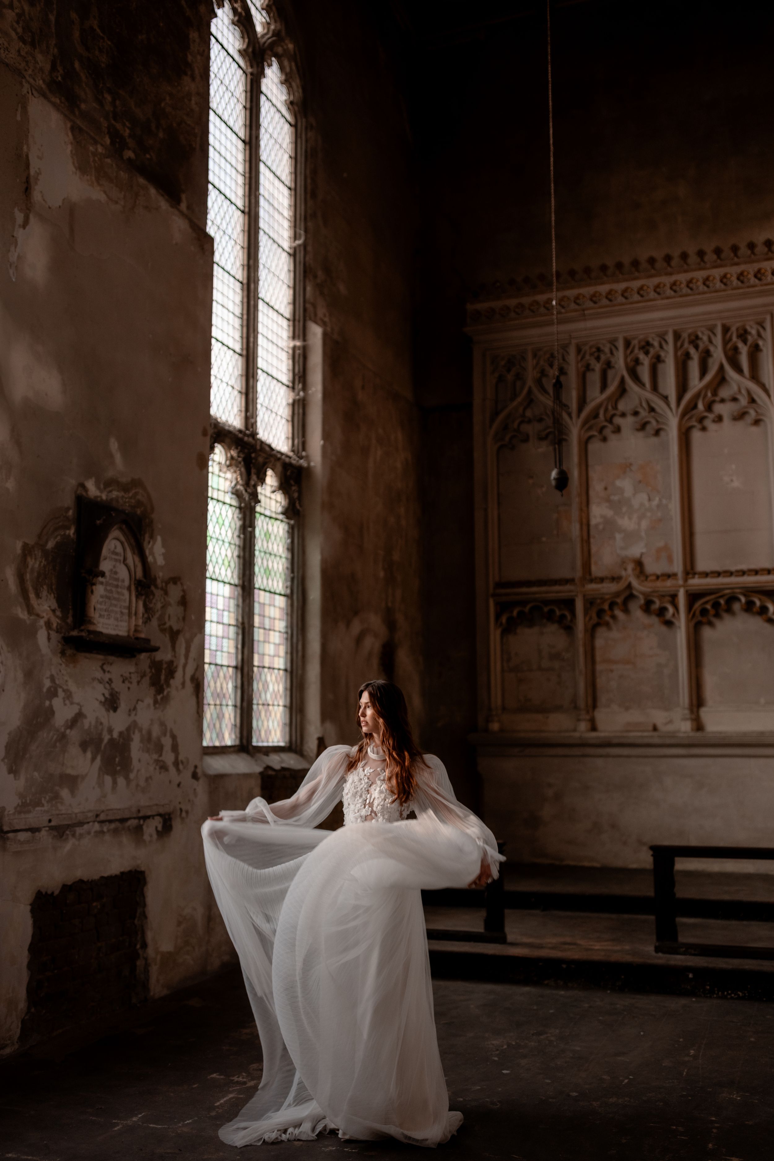 Bride in white lace gown poses in historic gothic chapel with tall windows and ornate stone architecture.